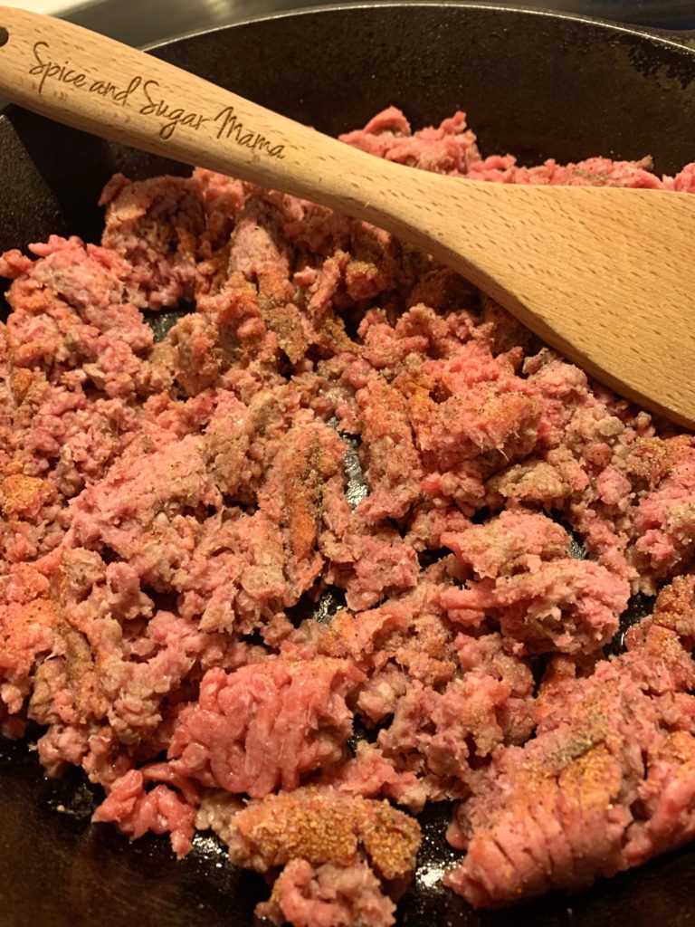 raw ground beef added to cast-iron skillet and being stirred with wooden spoon