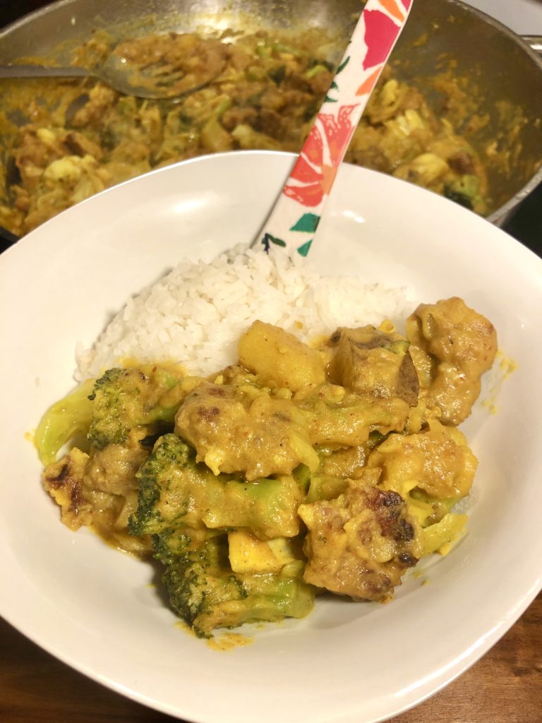 thai yellow curry with sausage and vegetables on white square bowl with rounded edges. photographed beside white rice with floral spoon. In the background is the large skillet with the rest of the dish.
