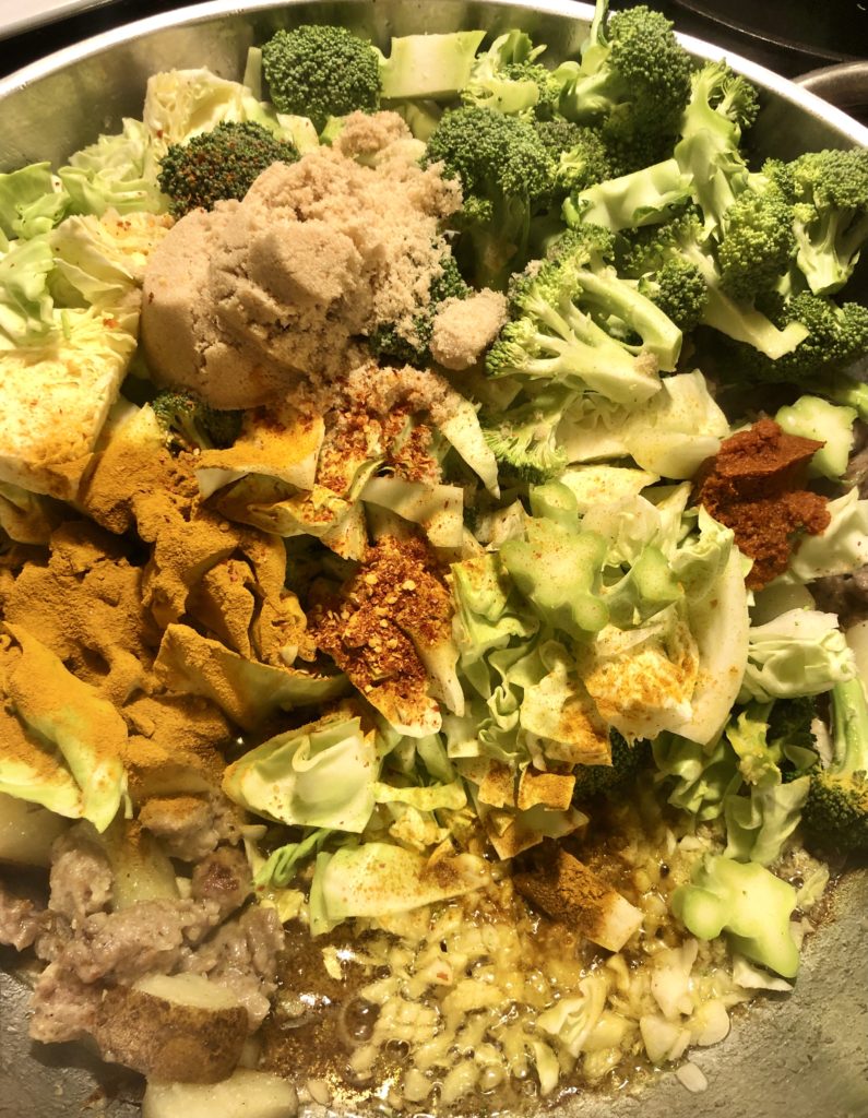 silver frying pan with sausage, potatoes, cabbage and broccoli. Turmeric, brown sugar, ground Thai chili peppers and red curry paste have been added to the pan.