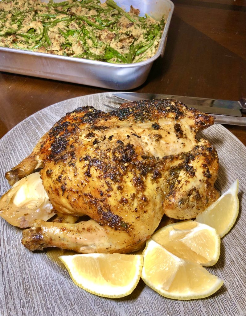Lemony oven roasted chicken being served on gray plate with lemon wedges pictured with green bean casserole in the background