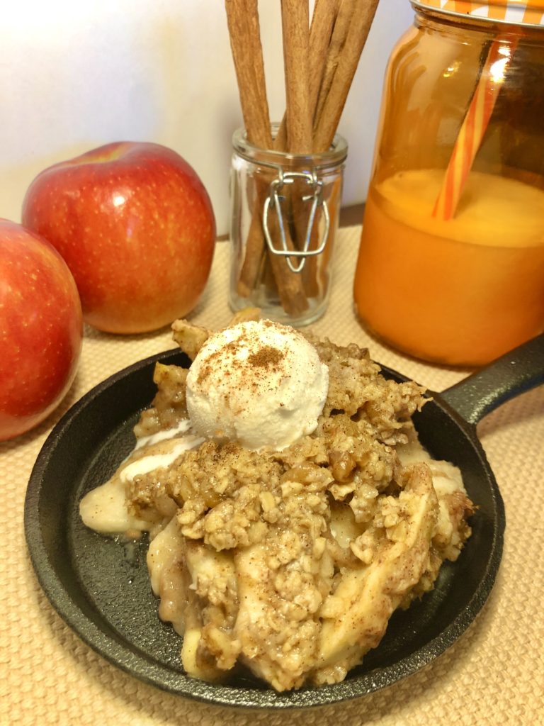 apple crisp topped with vanilla ice cream and a sprinkle of cinnamon in a mini cast iron skillet. skillet is photographed on tan fabric with two red apples, a jar with cinnamon sticks and an orange mason jar filled with milk in the background