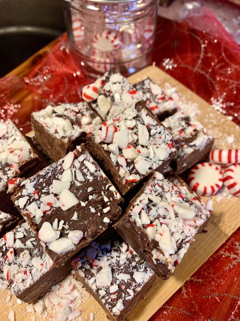 squares of peppermint fudge on oak cutting board pictured with starlight mint candies. These are on top of a red and silvery christmas fabric with a small jar filled with starlight mint peppermint candies