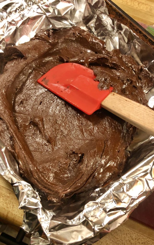 spreading prepared fudge in 8x8 pan so that it may set. The fudge is being spread over an aluminum foil lined pan and is being spread with a red rubber spatula