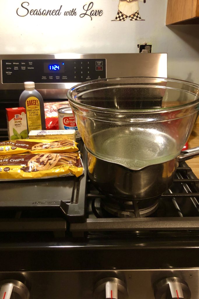 a makeshift double boiler made of saucepan and heat-proof glass bowl. The ingredients for the recipe are pictured on the stove. there are dark chocolate chips, vanilla extract, peppermint extract and salt