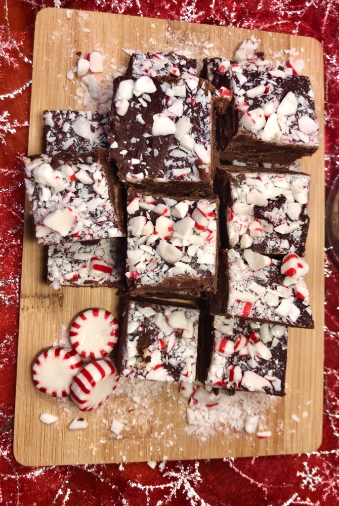 overhead shot of squares of peppermint fudge on oak cutting board. pictured with three whole starlight mint candies and broken up candies spread around the fudge. the cutting board is pictured on a red and silver fabric
