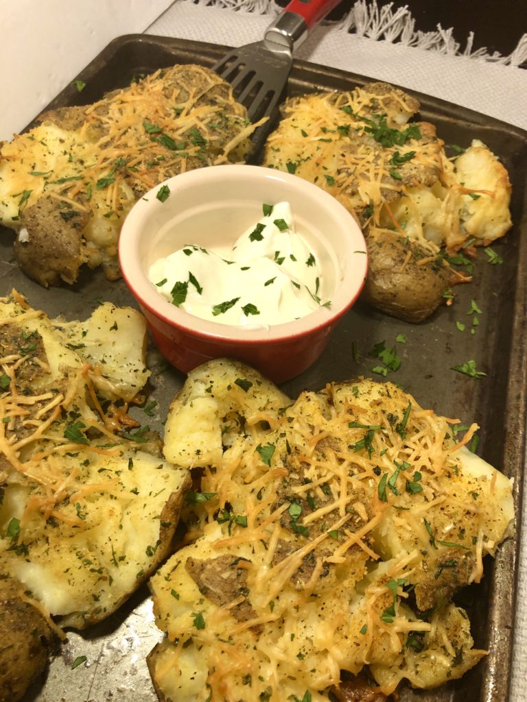 Four parmesan and olive oil smashed potatoes on a pan with a spatula under one potato and a red and white ramiken in the middle of the pan with sour cream. the entire pan and sour cream are garnished with fresh parsley