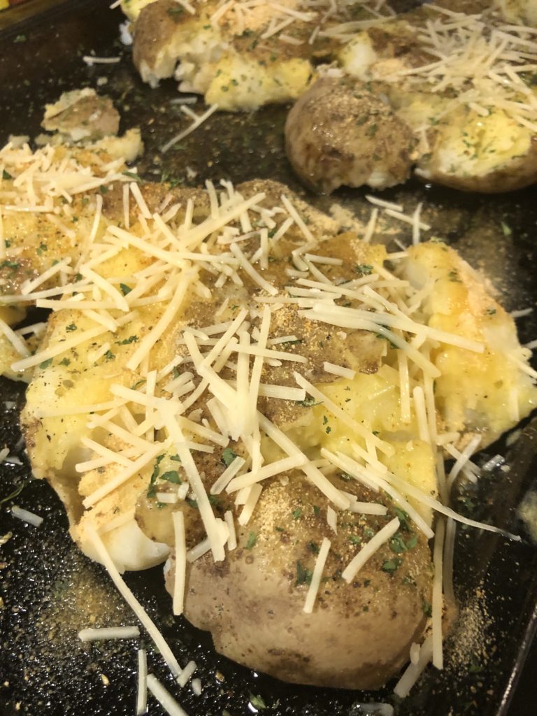smashed russet potatoes on pan with salt, black pepper, parsley, oregano, garlic powder, butter, olive oil and shredded parmesan cheese before baking in oven. The potatoes have been boiled whole and lightly smashed with a potato masher.