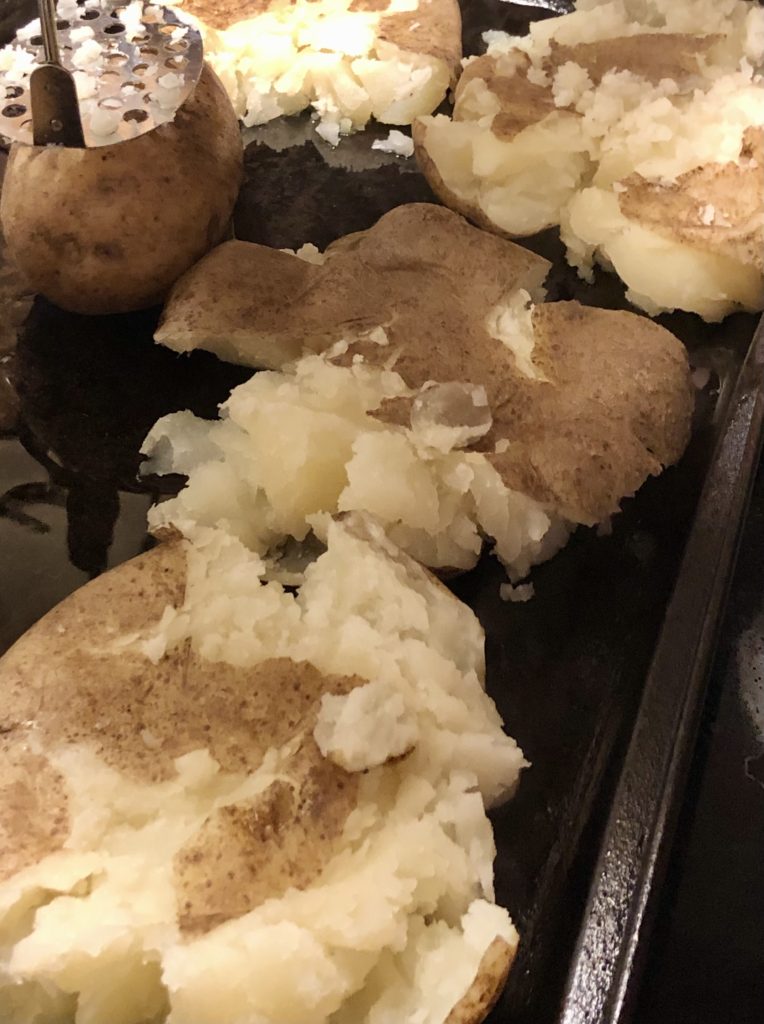 A baking pan of boiled russet potatoes that have been lightly smashed and pushed down with a hand masher.
