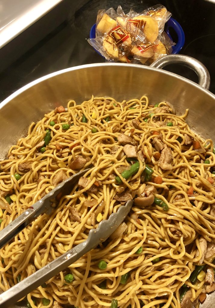 large silver skillet with chicken lo mein being tossed with tongs on stovetop. There is a blue bowl beside the skillet filled with Chinese fortune cookies