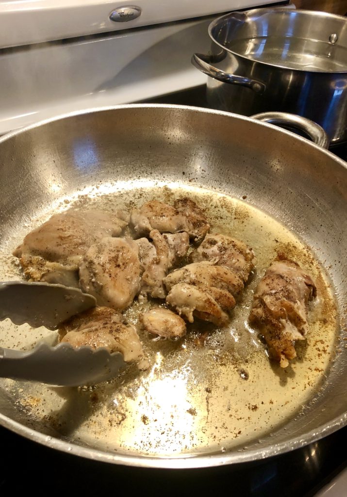 boneless skinless chicken thighs being fried in large silver skillet and being turned with tongs.