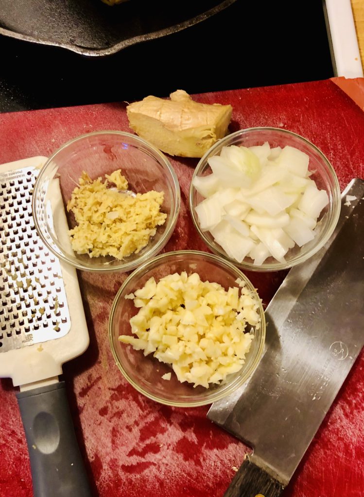 red cutting board with three clear ramikens. One filled with garlic, one ginger and one onion. There is a chopping knife and grater for the ginger on the cutting board as well.