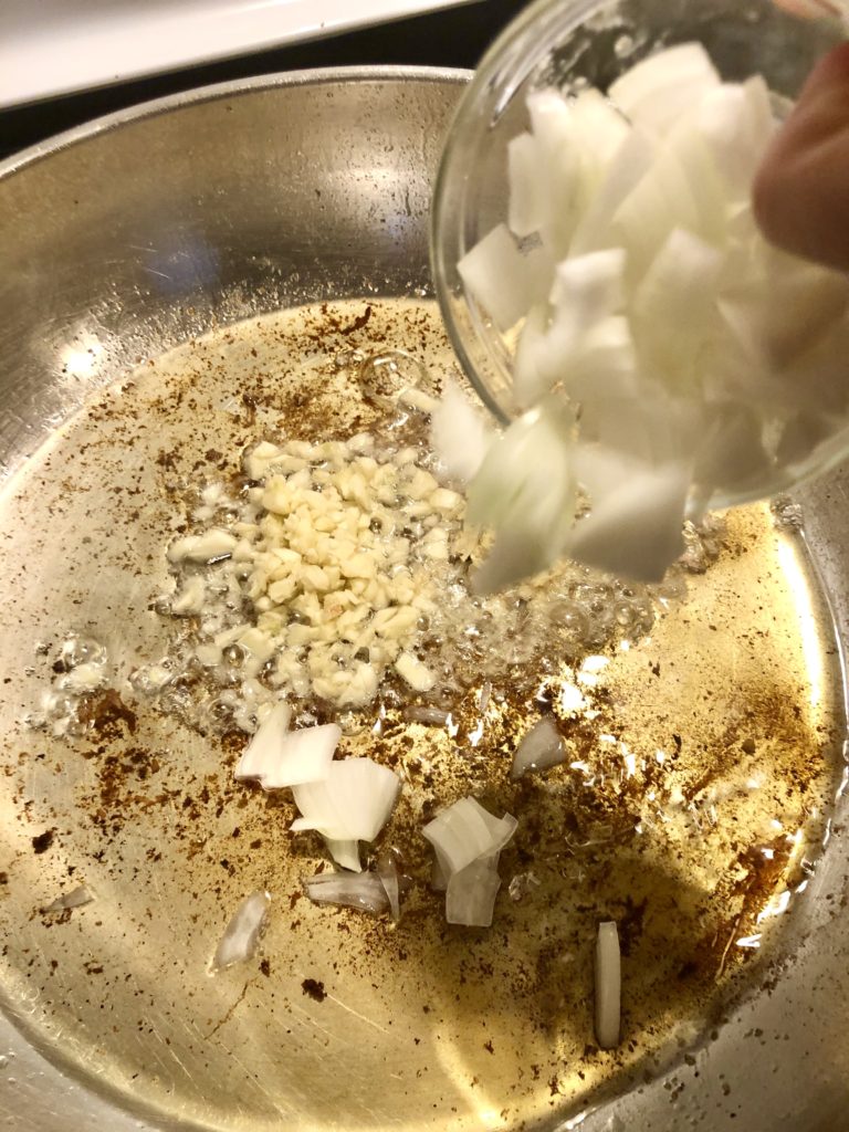 large skillet with vegetable oil and garlic, onion and giinger being added to the pan.