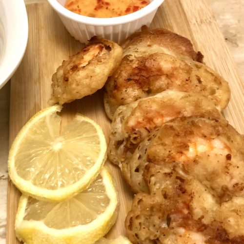 Six pieces of Chinese battered fried shrimp pictured on an oak cutting board alongside three lemon slices. There is a white bowl in the background with a bottled Thai chili dipping sauce