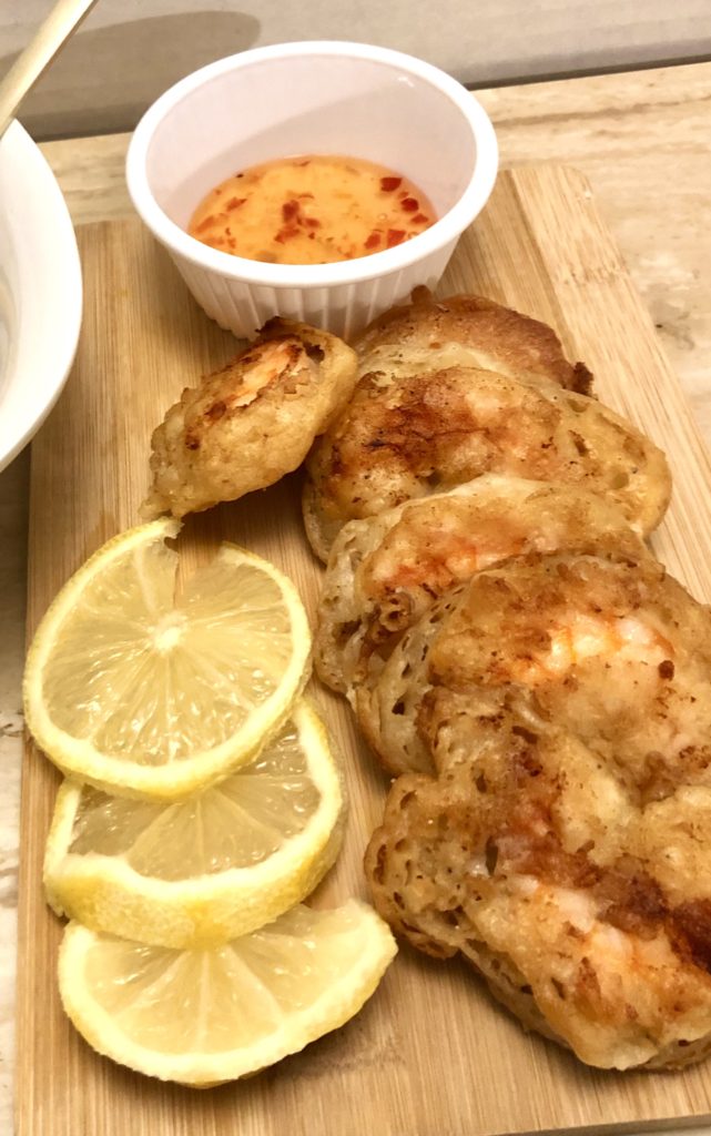 Six pieces of Chinese battered fried shrimp pictured on an oak cutting board alongside three lemon slices. There is a white bowl in the background with a bottled Thai chili dipping sauce