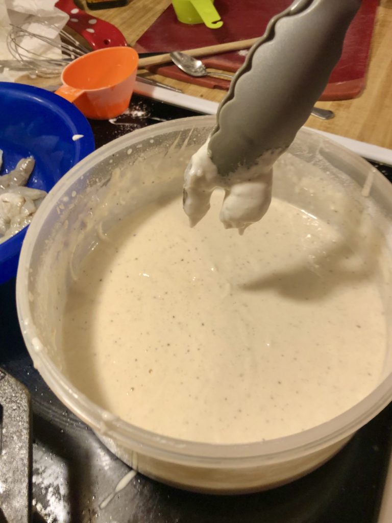 tongs holding a piece of shrimp over the batter for the excess to drip off. There is a blue bowl with shrimp in the background waiting to be dipped.