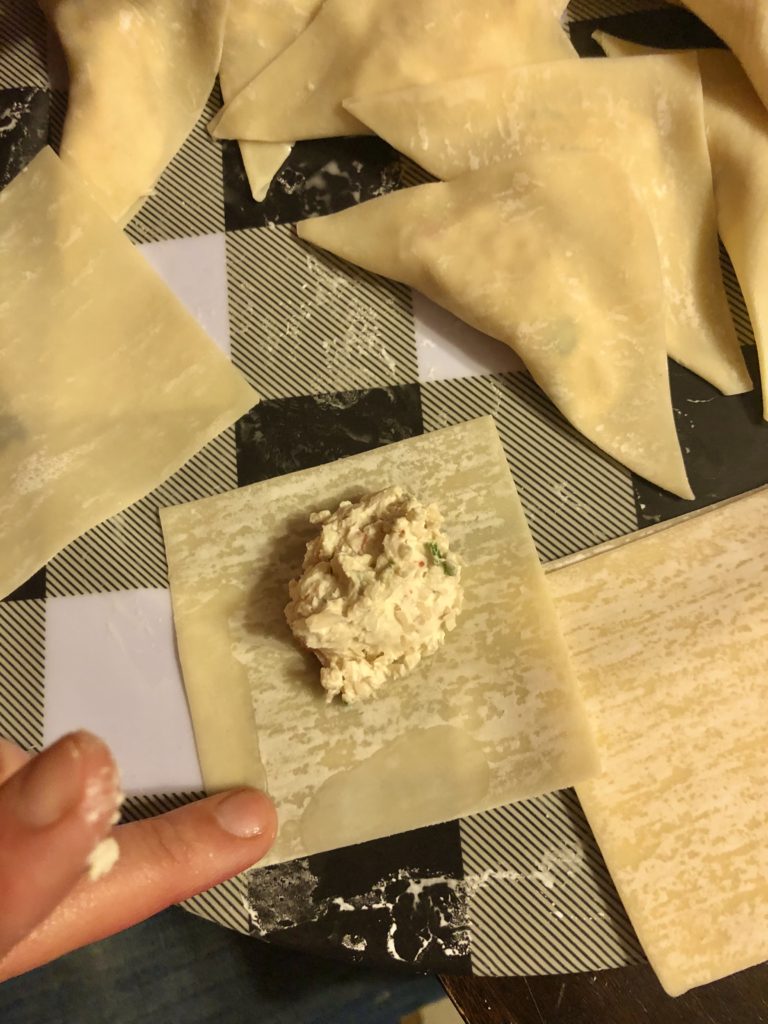 preparing crab rangoons on a black and white plaid plate. There is a prepared wonton wrapper with a tablespoon of the crab rangoon filling in the middle of the wrapper. a finger is adding warm water to the edges to prepare for folding over and sealing shut