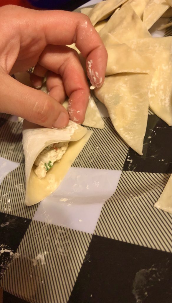 preparing crab rangoons. the filling has already been added and warm water has been placed on the edges and the wonton wrappers are being folded in this photo. There are other crab rangoons in the background and they are all prepped on a black and white plaid plate