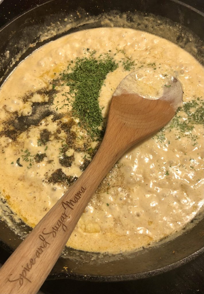 cajun shrimp pasta sauce being cooked in cast iron skillet. garlic, butter, cream, parm cheese, lemon zest, salt, black pepper, garlic powder, dried parsley, pasta water and mozzarella cheese are being stirred together with a wooden spoon.