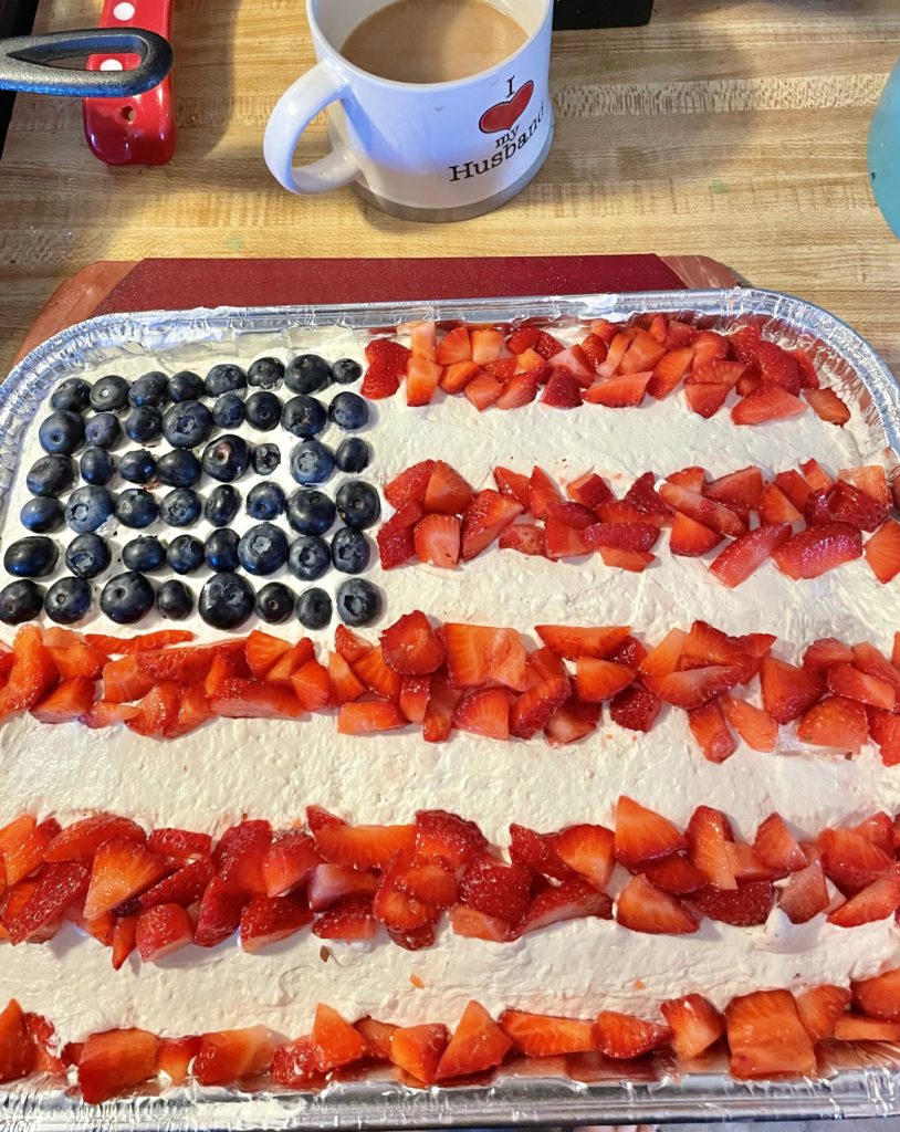 Strawberry cake with cream cheese and cool whip frosting. The top of the cake is designed with blueberries and strawberries that are arranged to resemble a flag. The cake is pictured in a tin foil dish on a red cutting board.