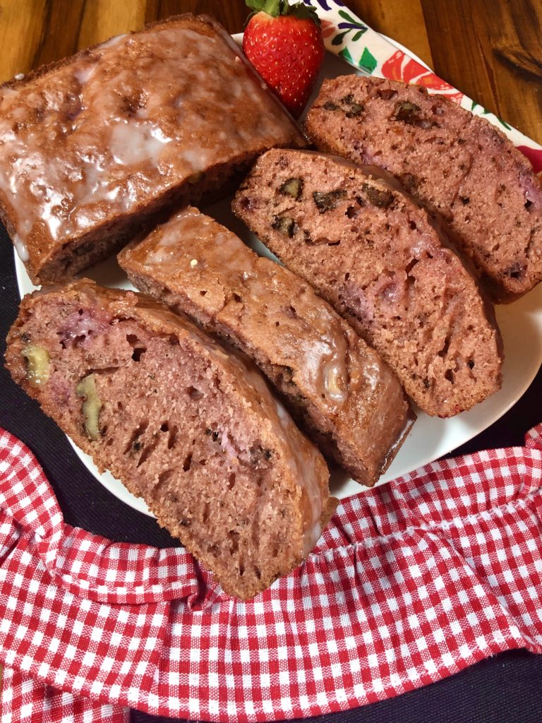 Four slices and a half loaf of strawberry walnut bread with lemon glaze on white square plate. Pictured with one strawberry and floral butter knife on top of wooden cutting board covered with white and red checkered fabric