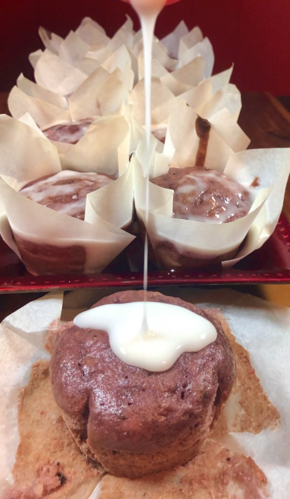 red plate with lemon glazed strawberry bread muffins. One unwrapped muffin is pictured in front of the plate with lemon glaze being drizzled over the top