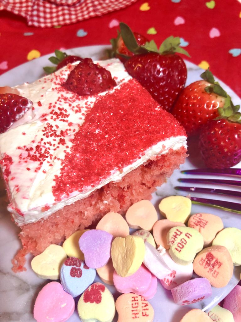 slice of strawberry cake with cream cheese and cool whip frosting. The cake is topped with fresh heart shaped strawberries and red sugar sprinkles. Played on white plate with purple fork. There are three fresh whole strawberries on the plate with scattered Sweetheart candies with words on them on the plate. There is red fabric with multi-colored hearts on the table in the background