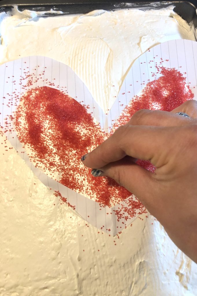 red sugar sprinkles being added to the inside of a paper heart cutout over cake frosting.