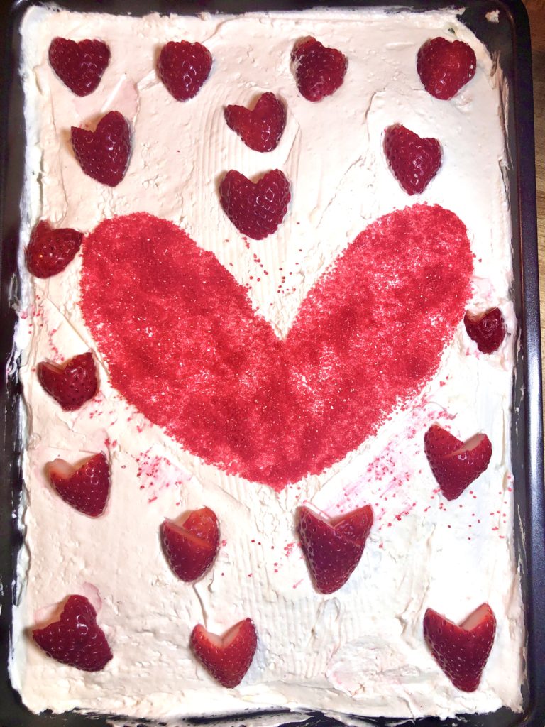 strawberry cake with white cream cheese and cool whip frosting in 13x9 sheet pan. The frosting is decorated with a red sugar sprinkle heart in the middle of the cake and the sprinkle heart is surrounded by fresh strawberries cut into the shapes of smaller hearts