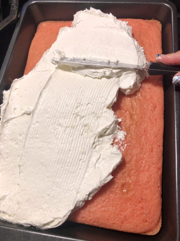 cream cheese and cool whip frosting being spread over baked strawberry sheet cake