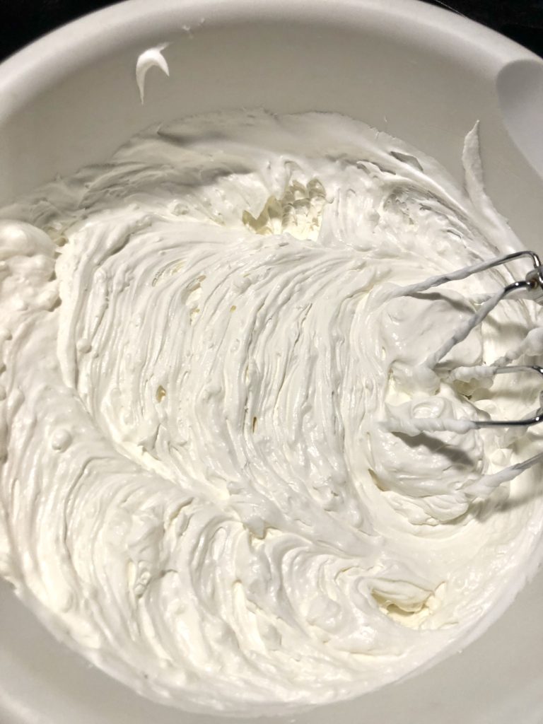 cream cheese cool whip frosting in white mixing bowl being whipped with electric mixer