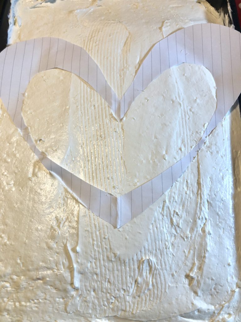 paper cutout of heart shape being pressed gently into the frosting of a 13x9 sheet cake