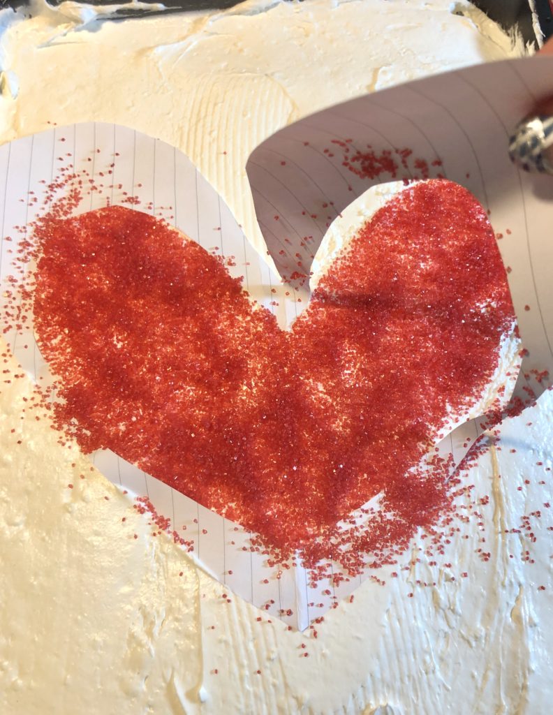 paper cutout of heart shape is being removed from cake frosting leaving a red sugar sprinkle heart shape in the middle of a cake with white cream cheese and cool whip frosting