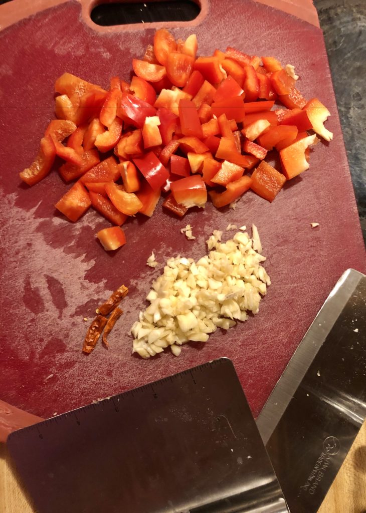 red cutting board with chopping knife lying beside it. Garlic, Thai chili peppers and red bell peppers are chopped on top.