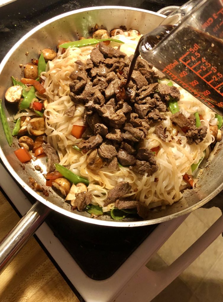 Rice noodles, cooked beef, stir-fry sauce being added to large skillet with cooking mushroom, red peppers and snow peas