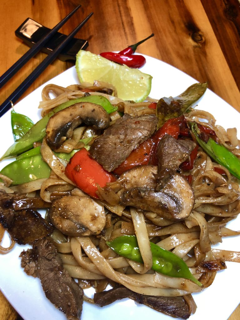 Thai beef rice noodles with mushrooms, snow peas and red bell peppers pictured on white square plate with lime wedge and two Thai chili peppers. Black chop sticks are pictured in the background and everything is on a wooden cutting board