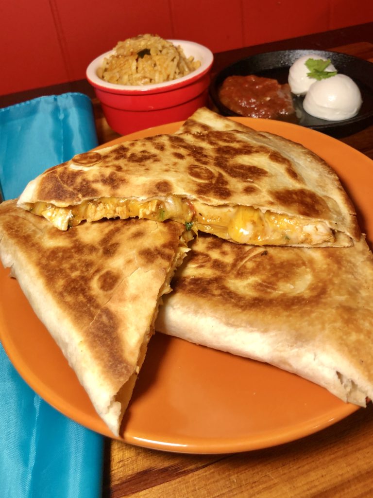 Three servings of cheesy chicken quesadillas with salsa arranged on an orange plate. There is a bright blue napkin pictured next to the plate and everything is on a wooden cutting board. In the background, there is a red ramekin with Spanish rice and beside that is a mini black cast iron skillet with salsa and sour cream for dipping. 