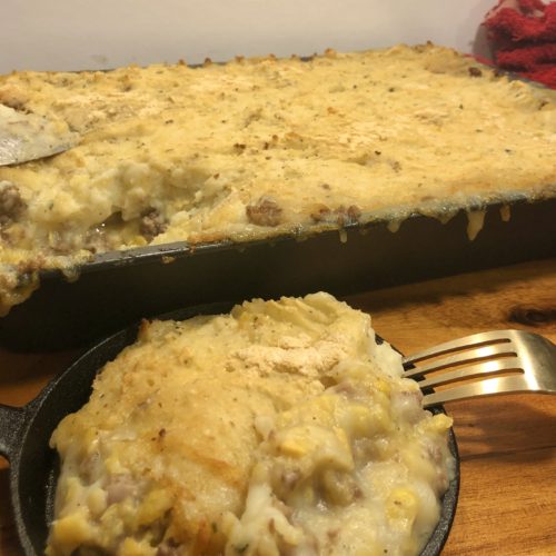 shepherd's pie is pictured in a miniature cast iron skillet with a fork. There is a 13x9 baking pan in the background of the photo of the shepherd's pie with a serving spoon next to it. Mashed potato from the dish is dripping down the side of the baking pan and both items are on top of a wooden cutting board.