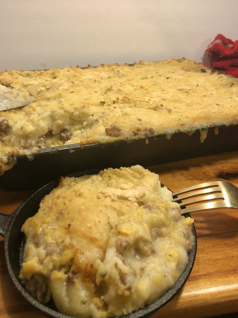 shepherd's pie is pictured in a miniature cast iron skillet with a fork. There is a 13x9 baking pan in the background of the photo of the shepherd's pie with a serving spoon next to it. Mashed potato from the dish is dripping down the side of the baking pan and both items are on top of a wooden cutting board.