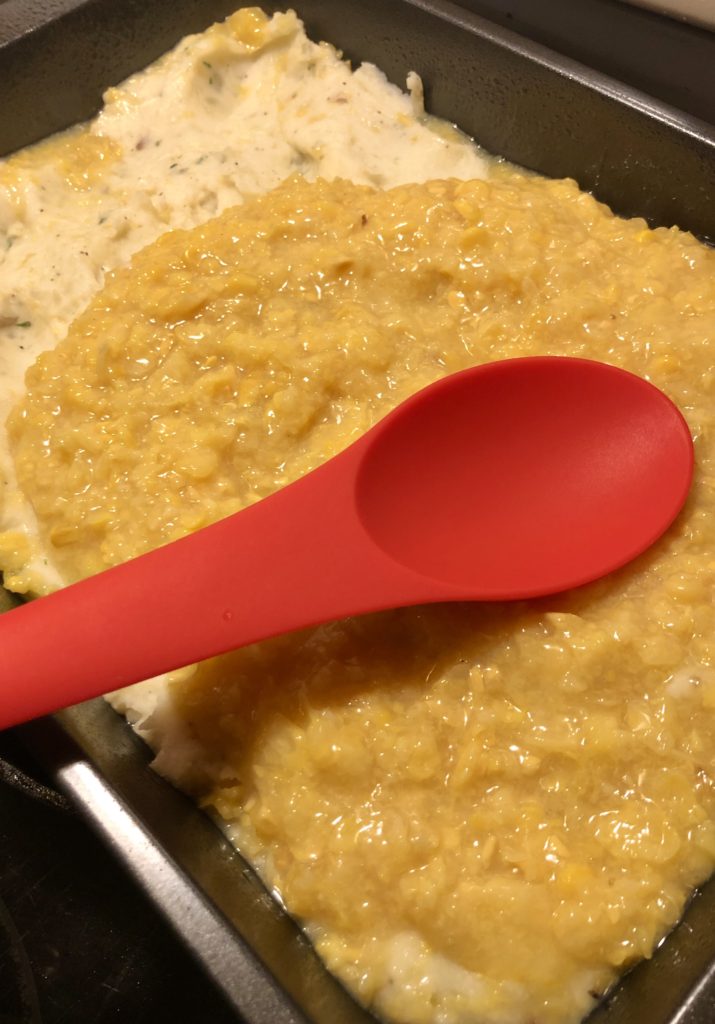 Canned cream corn being spread over layer of mashed potatoes in 13x9 baking dish. The corn is being spread into an even layer with a red spoon.