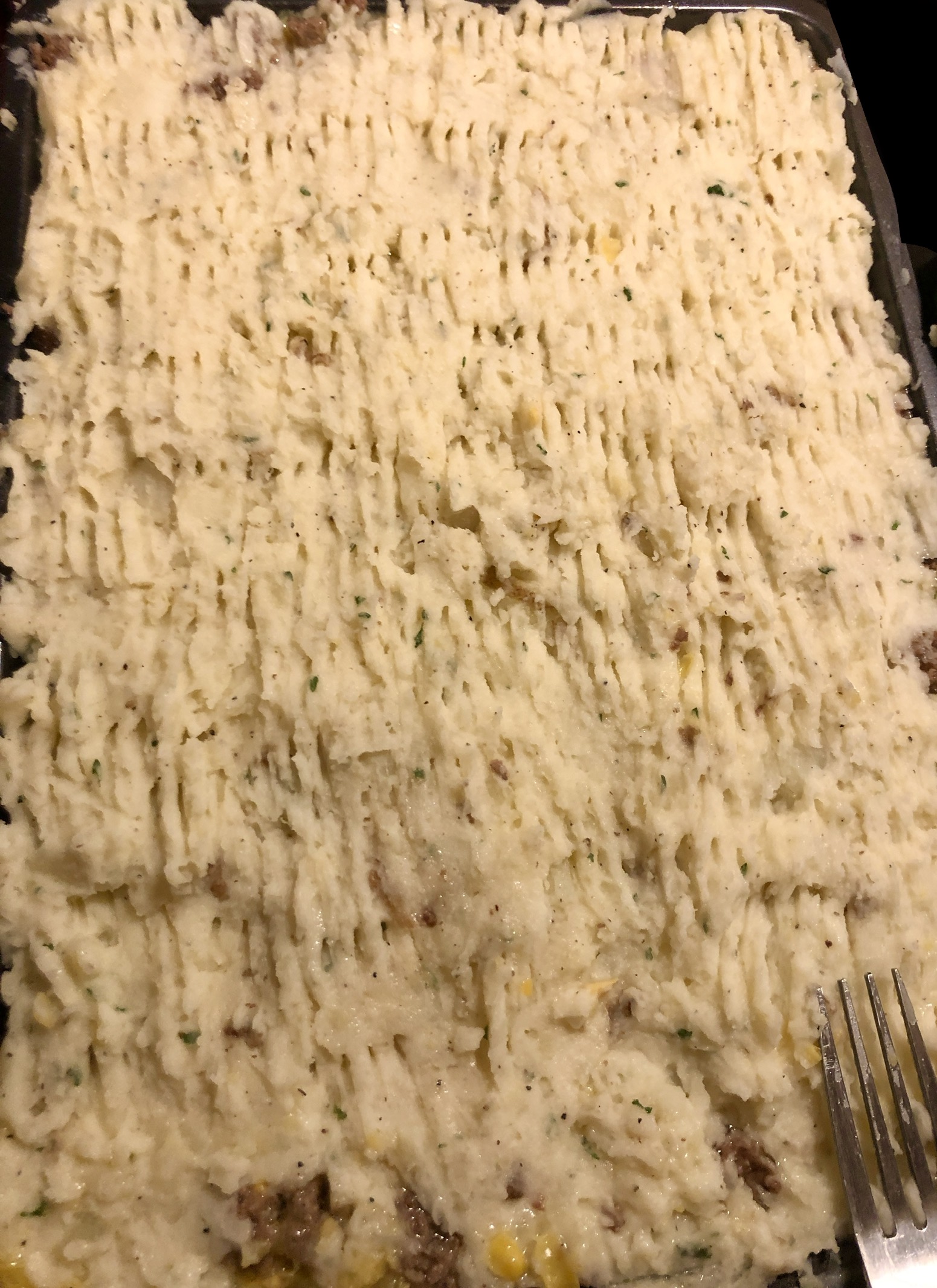 Top layer of mashed potatoes over a shepherd's pie that is in a baking dish. A fork has been used to make a rake pattern over the top of the mashed potatoes.
