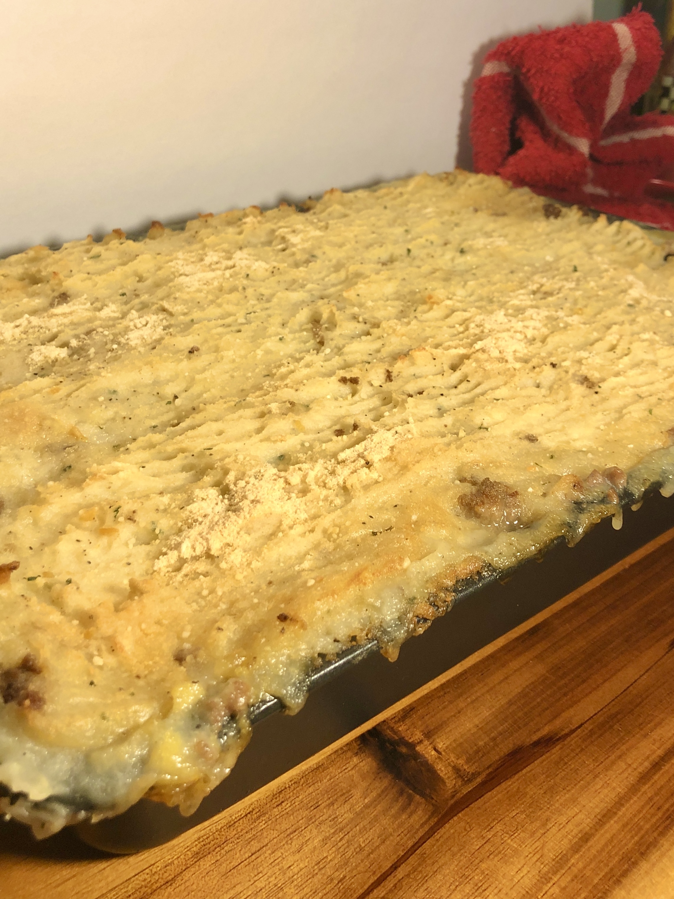 Side view of a baked Shepherd's Pie in 13x9 baking pan. The pan is cooling on a wooden cutting board with a red towel off in the background.