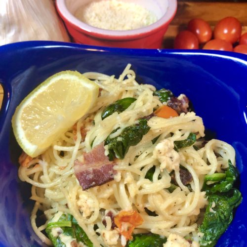 Spinach, bacon, tomato, feta fettuccine plated in royal blue bowl with a lemon wedge. In the background is a whole garlic bulb, a ramiken with grated parmesan cheese and cherry tomatoes.