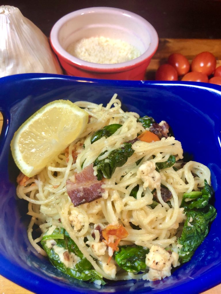 Spinach, bacon, tomato, feta fettuccine plated in royal blue bowl with a lemon wedge. In the background is a whole garlic bulb, a ramiken with grated parmesan cheese and cherry tomatoes. 