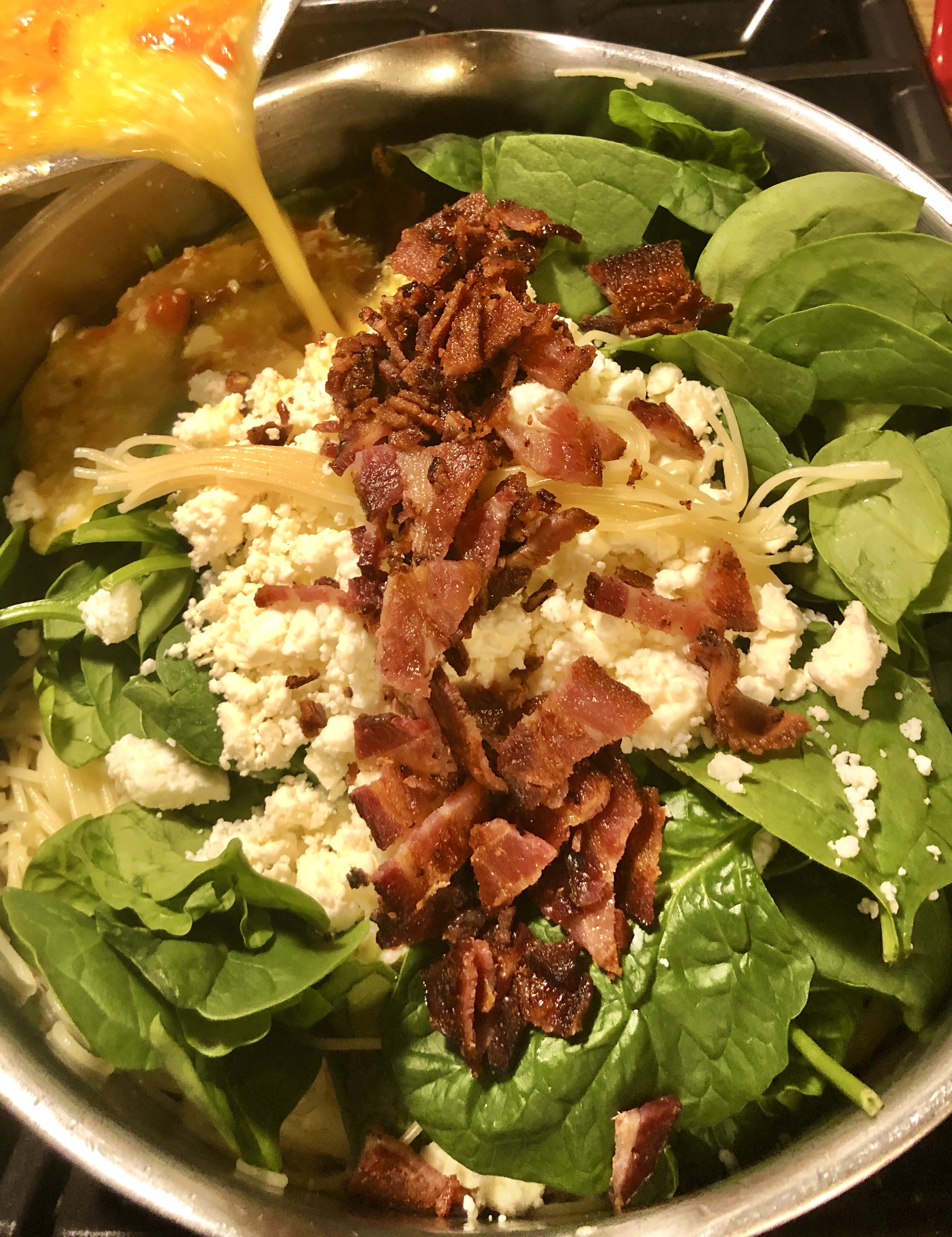 Bacon, spinach and feta over fettuccine in a pot. Lemon parmesan sauce is being poured over the top.