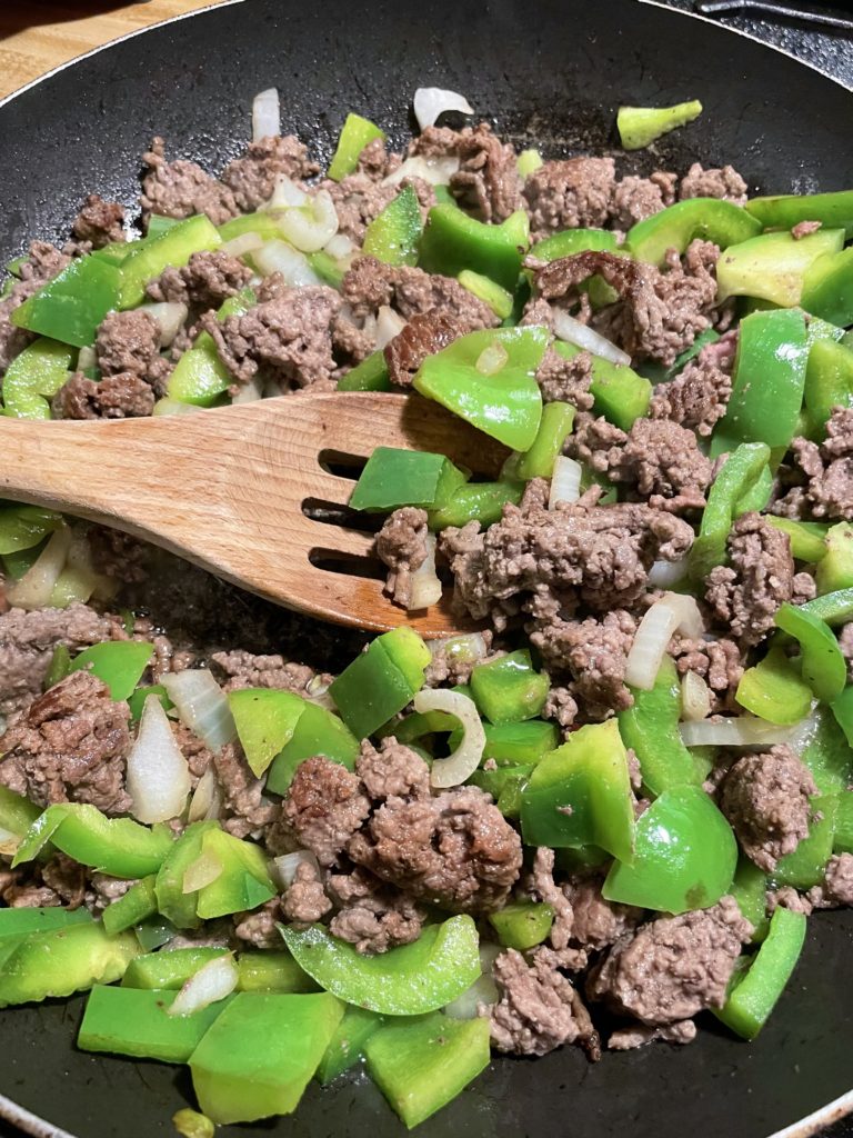Ground beef, green bell peppers, onion and garlic frying together in a skillet and being stirred by a wooden spoon.