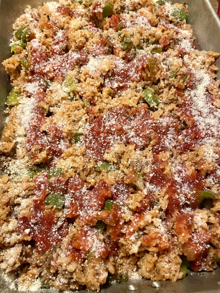 Unstuffed pepper rice casserole pictured with pasta sauce and grated parmesan cheese over the top prior to baking in the oven. The food is pictured in a 13x9 baking pan.