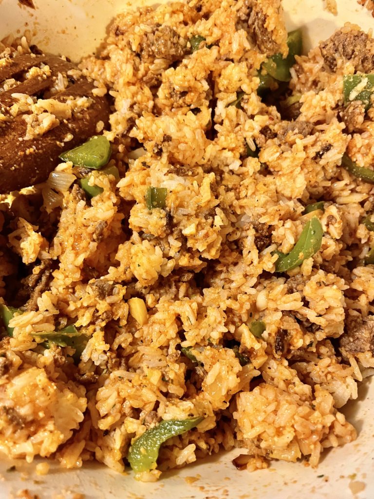 Rice, ground beef, green pepper and pasta sauce being stirred together in a large white mixing bowl with a wooden spoon.
