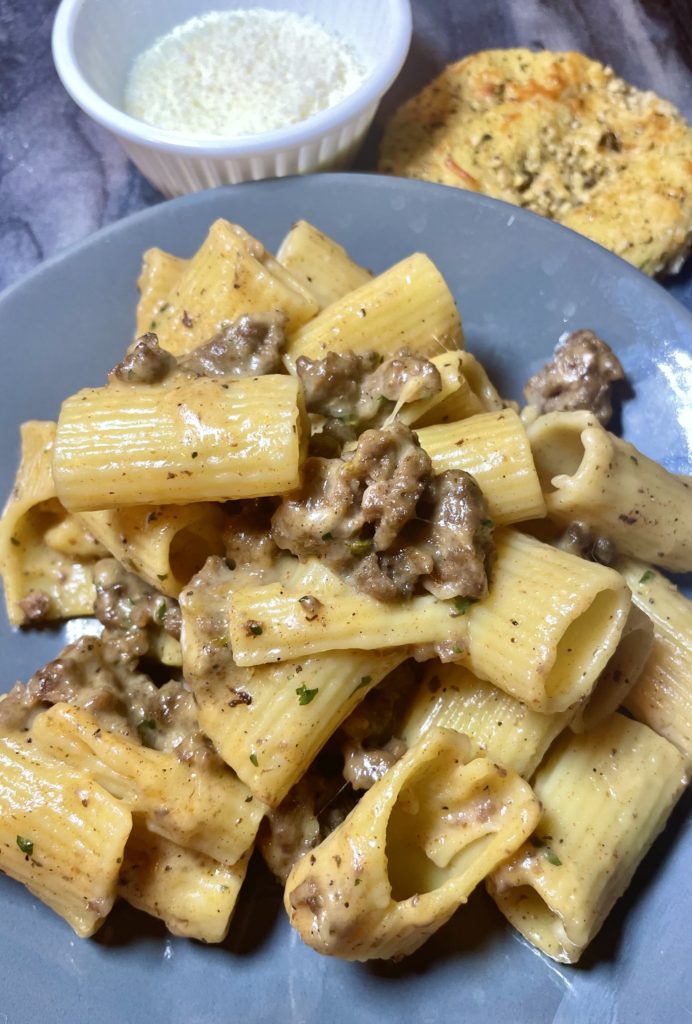 Homemade Hamburger Helper made with rigatoni pasta and a cheese sauce with ground beef. Pasta is plated on a gray plate with a white ramekin of grated parmesan cheese and a round slice of garlic bread in the background.