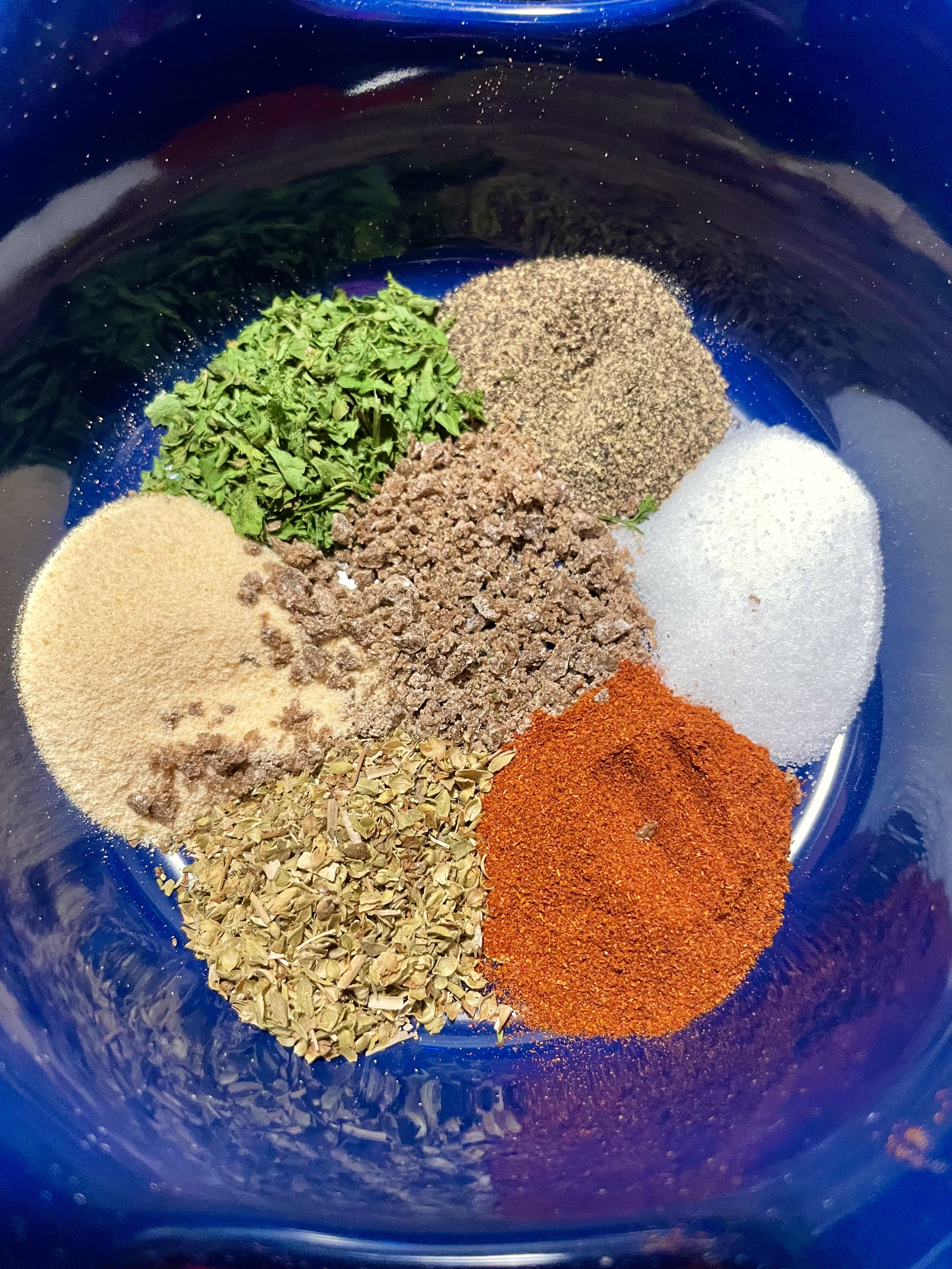 A blue bowl with salt, black pepper, beef bouillon powder, paprika, dried parsley, dried oregano and garlic powder.