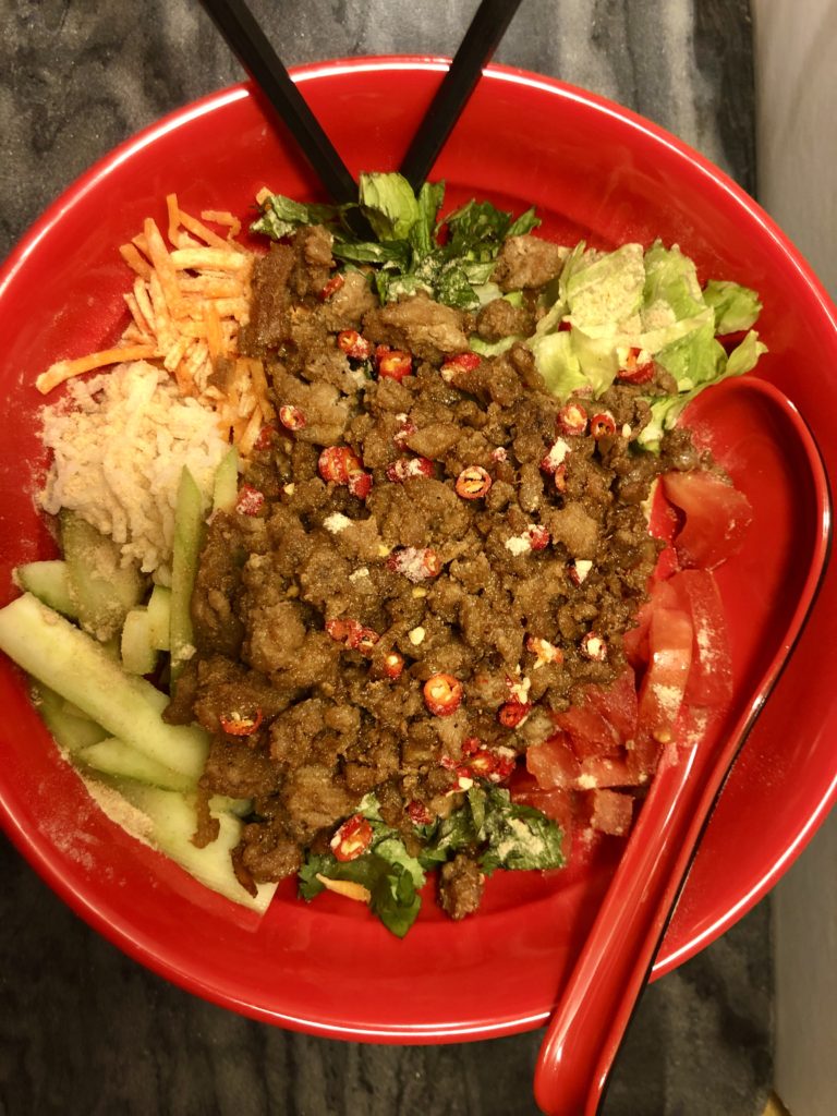 Thai caramelized ground pork in a Jasmine rice bowl. The rice bowl contains lettuce, cucumber, carrots, cilantro, mint, Thai chilis and toasted rice powder. Pictured in a red bowl with a red soup spoon and black chopsticks on top of a charcoal marbled table.
